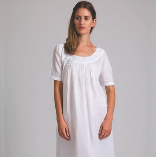 A woman standing in a white, short sleeve, cotton nightie with a scoop neckline and ruffled hem.